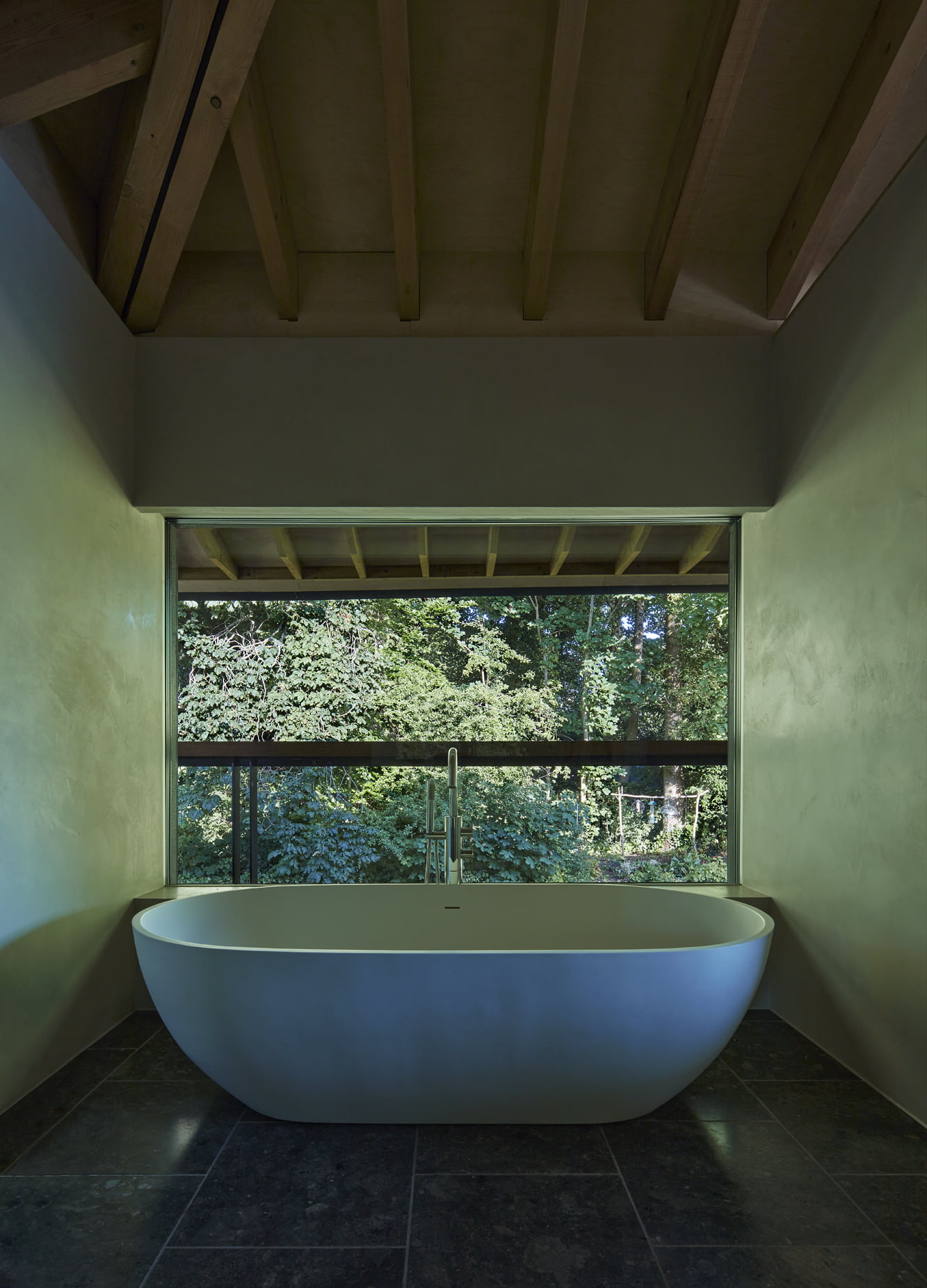 A contemporary bathroom with a free-standing bath tub at the end of it, looking out of a large window onto a woodland area.