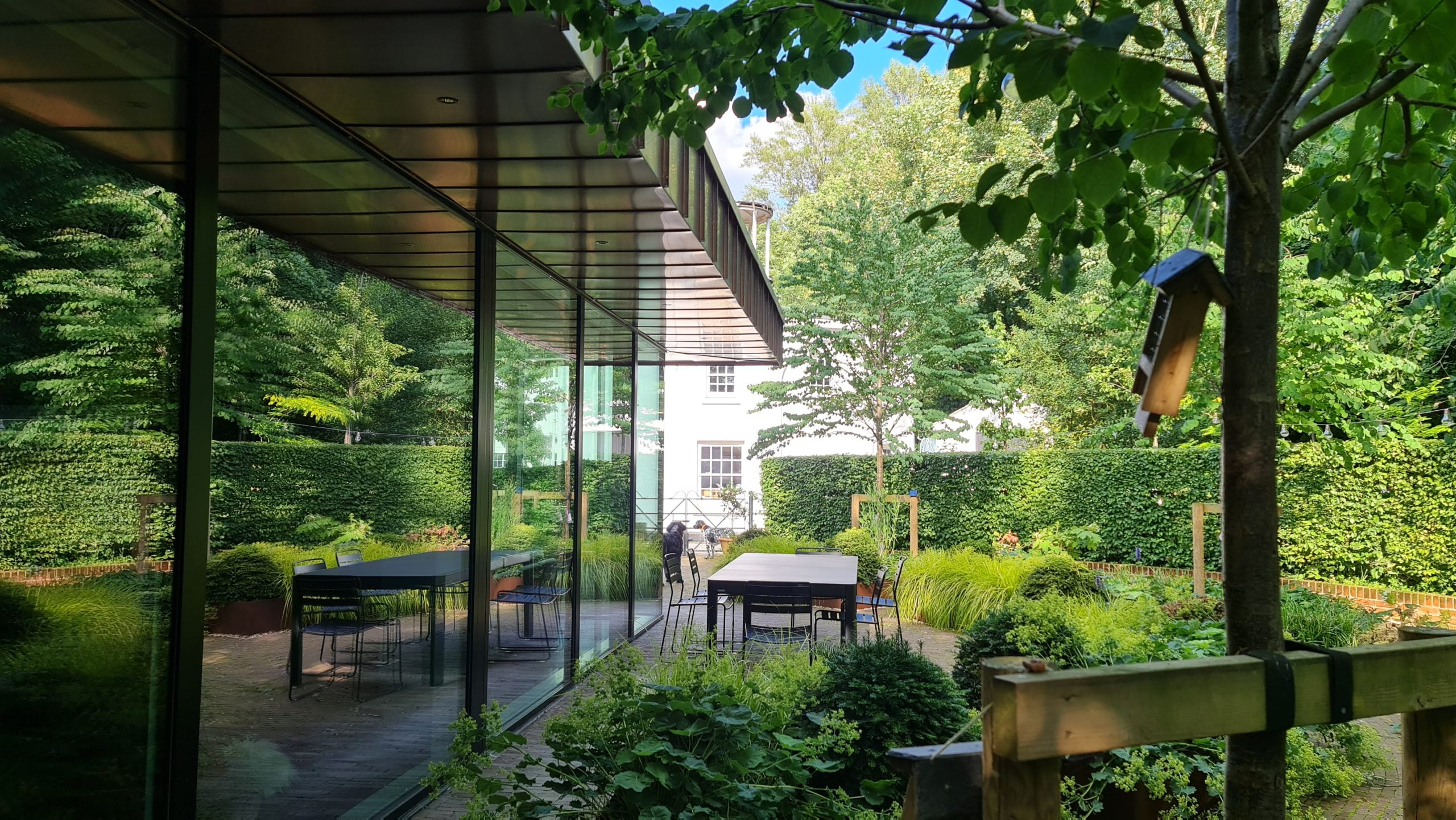 The patio of a modern house, surrounded by greenery and tress with a table and chairs in the middle of it.