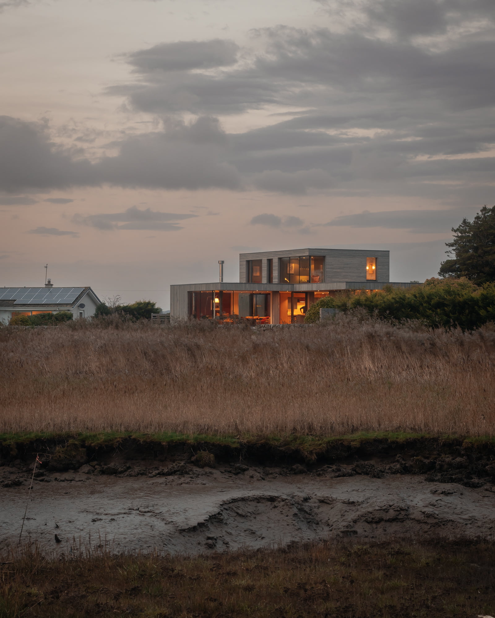 A field with long grass and a modern house in the background with the lights on.