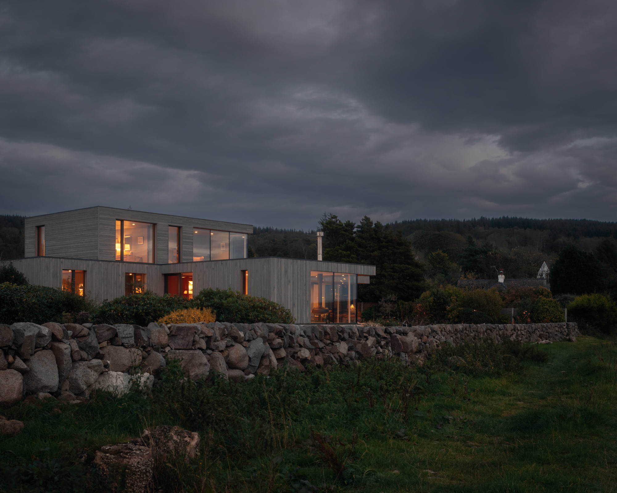 A contemporary wooden-cladded property with large, floor to ceiling windows and a stone wall surrounding it.